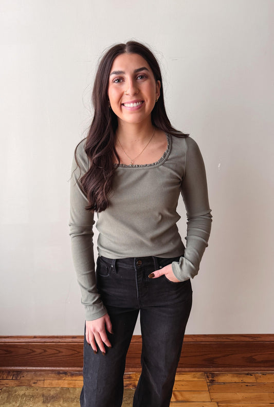 A woman with long dark hair stands indoors wearing a fitted long-sleeve top in a muted sage green color with dark charcoal jeans, posing against a plain off-white wall with a wooden baseboard and floor.