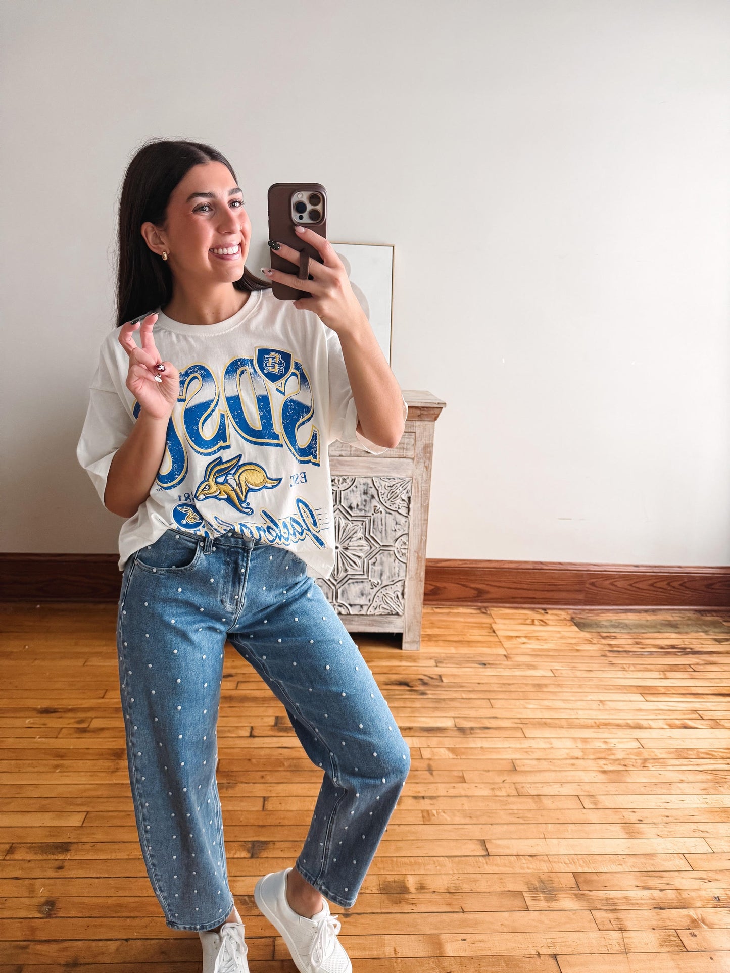 A woman takes a mirror selfie in a room with light wood floors and a white wall. She is wearing a white graphic t-shirt with blue and yellow 'Beast' lettering, blue jeans with small white polka dots, and white sneakers.