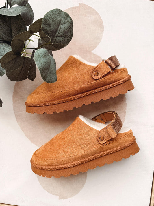 A pair of tan suede slip-on clogs with white shearling lining and a brown strap, displayed on a light beige background with a decorative plant leaf in the corner.