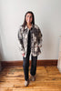 A woman stands in a room with a light gray wall and wooden floor, wearing a gray and black mottled camouflage jacket over a black top and black pants.