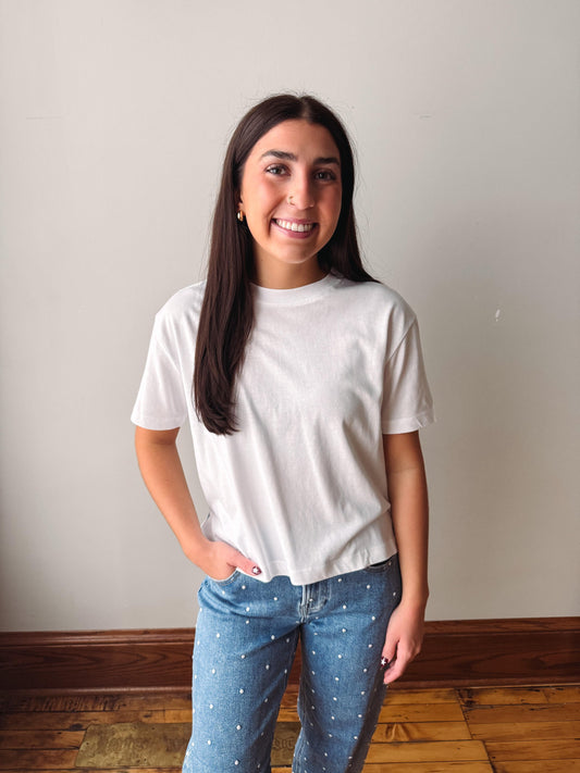 A woman with long dark hair smiling, wearing a white short-sleeve crewneck t-shirt and blue jeans with small white star patterns, standing against a plain light gray wall with a wooden floor.