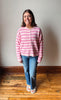 A woman standing in a room with a light gray wall and wooden floor, wearing a pink and white striped fuzzy cardigan, blue jeans, and white slide sandals.
