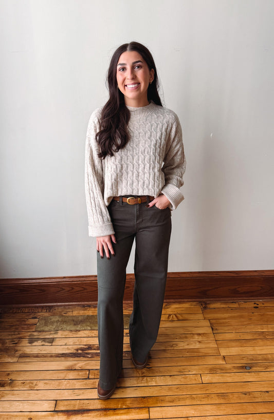 A woman stands in a room with a light gray wall and wooden floor, wearing a cream cable-knit sweater, dark gray wide-leg pants, and brown boots.