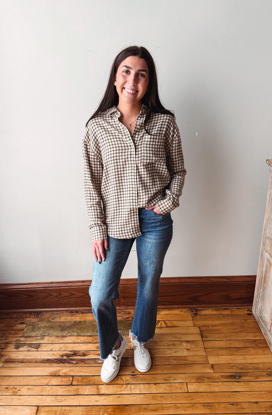 A woman stands in a room with light-colored walls and wooden floors, wearing a brown and beige gingham long-sleeve button-up shirt, blue jeans, and white sneakers.