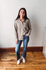 A woman stands in a room with light-colored walls and wooden floors, wearing a brown and beige gingham long-sleeve button-up shirt, blue jeans, and white sneakers.