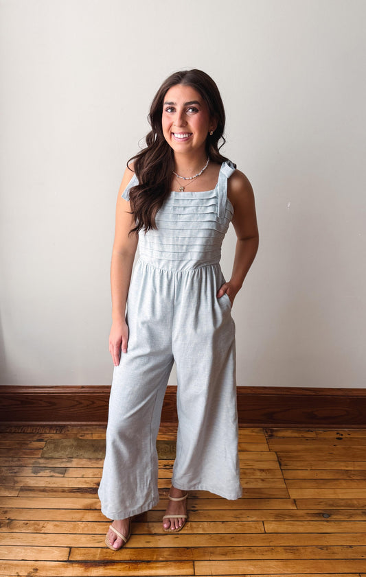 Woman wearing a light blue jumpsuit standing on a wooden floor with a white wall background