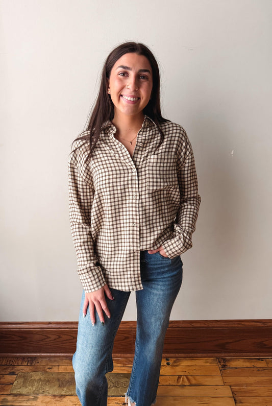A woman with long dark hair stands smiling, wearing a brown and beige gingham button-up shirt and blue jeans, posing in front of a plain off-white wall with a wooden baseboard and hardwood floor.