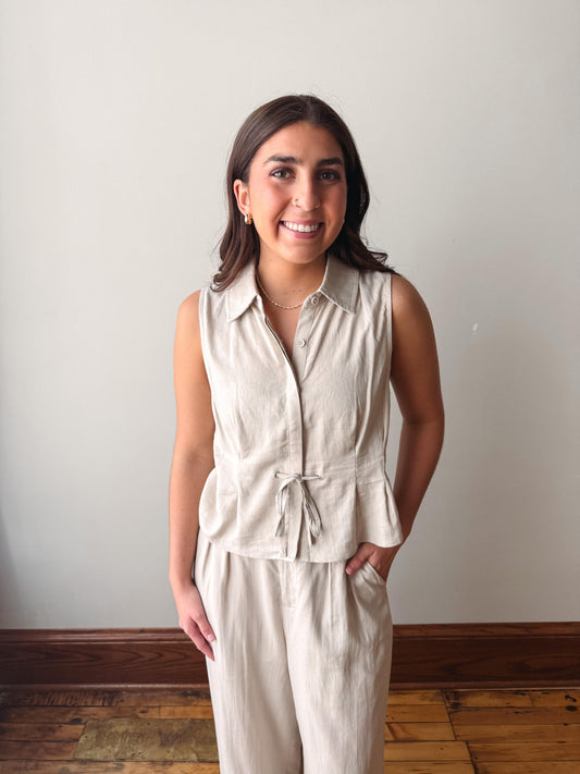 Woman wearing a beige sleeveless top and pants against a plain wall.