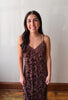 A woman with long dark hair stands indoors wearing a sleeveless, V-neck slip dress with a dark floral lace pattern over a muted brown base. She is posing in front of a plain off-white wall with a wooden baseboard and hardwood floor visible.