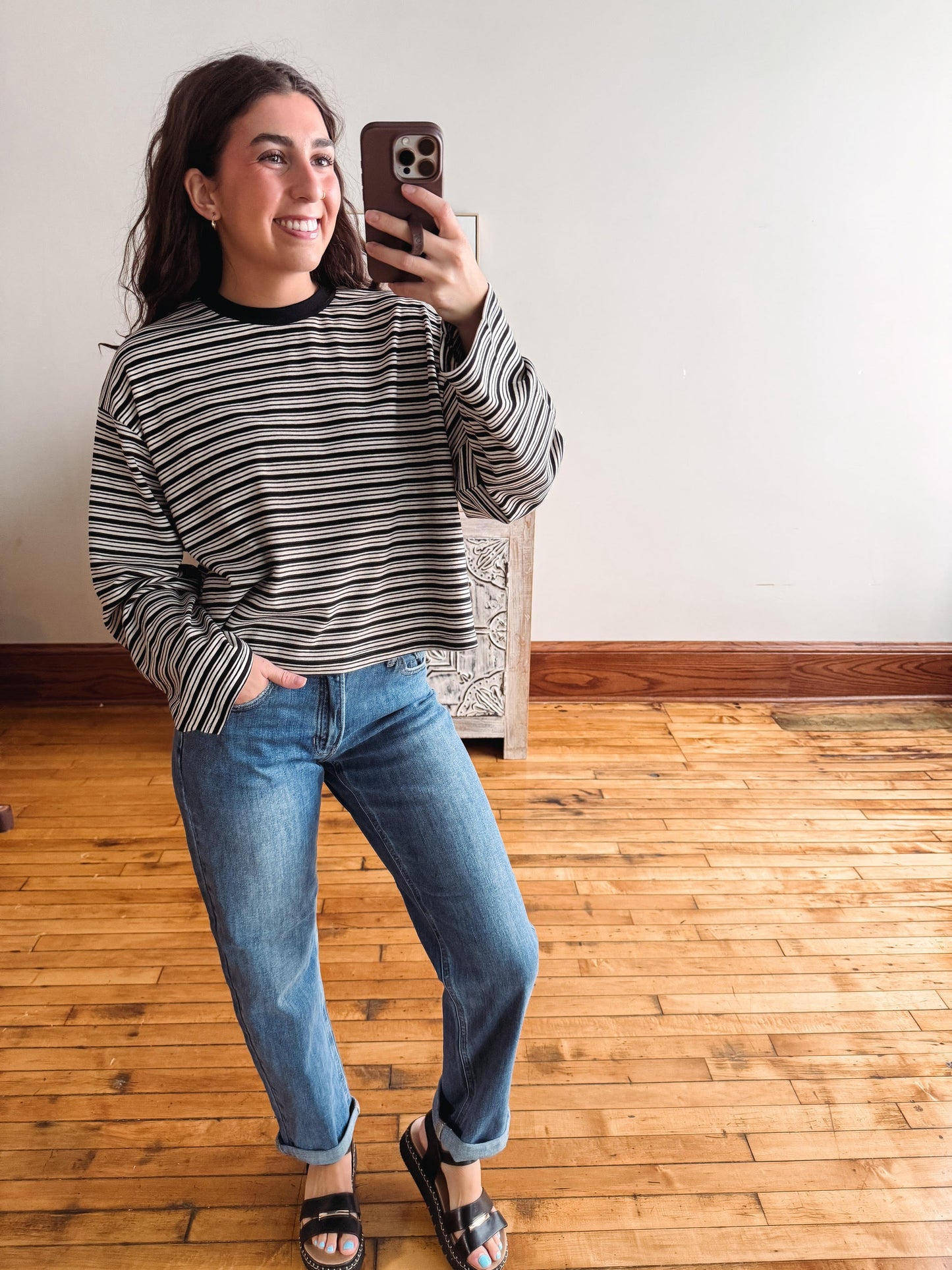 A woman takes a mirror selfie wearing a black and white horizontally striped long-sleeve shirt, blue jeans, and black slide sandals in a room with a light-colored wall and wooden floor.