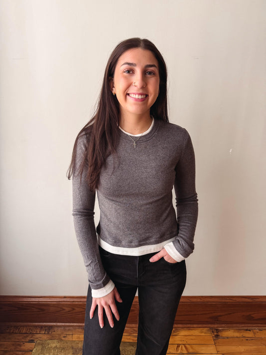 A woman smiling while wearing a dark gray long-sleeve crewneck sweater with white ribbed trim at the collar, cuffs, and hem, paired with black pants, standing against a plain beige wall.
