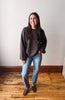 A woman stands in a room with a light gray wall and wooden floor, wearing a dark brown knit sweater, blue jeans, and brown suede loafers.
