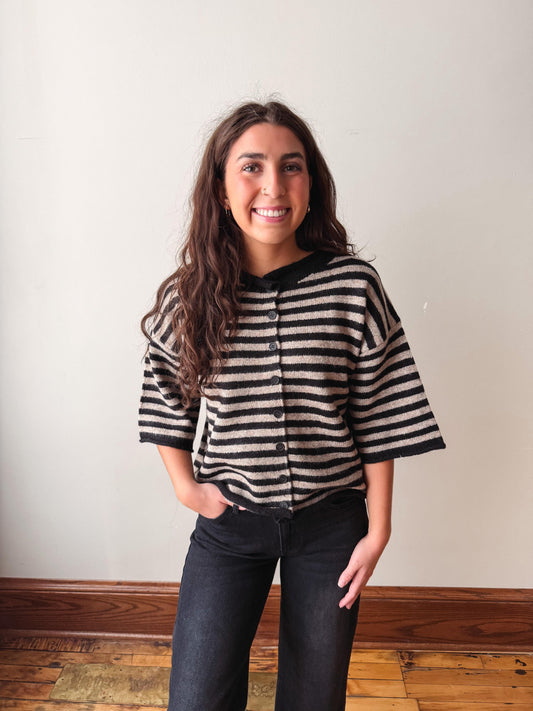 A smiling young woman with long wavy brown hair stands indoors wearing a black and beige horizontally striped short-sleeve button-front cardigan or sweater with dark jeans, against a plain off-white wall and wooden floor.