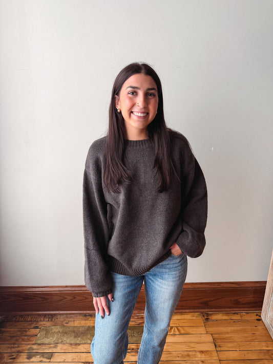 A woman with long dark hair stands indoors wearing a dark brown oversized knit sweater and light blue jeans, with a plain off-white wall and wooden floor behind her.