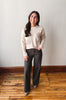A woman modeling a cream knit sweater with brown floral lace-up details on the sleeves, paired with dark gray wide-leg pants and brown boots, standing on a wooden floor against a plain wall.