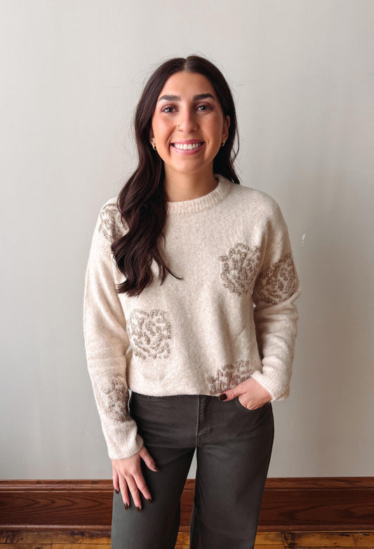 A smiling woman with long dark hair stands indoors wearing a cream knit sweater with taupe floral motifs and dark charcoal pants against a plain light wall.