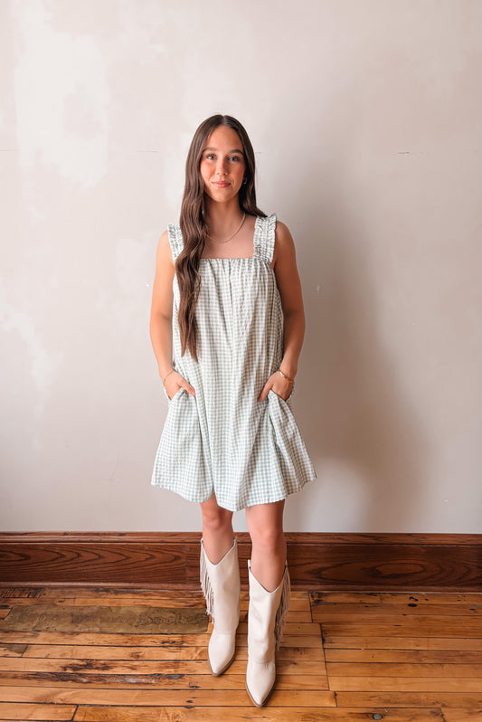 Woman wearing a checkered dress and white boots standing against a plain wall.
