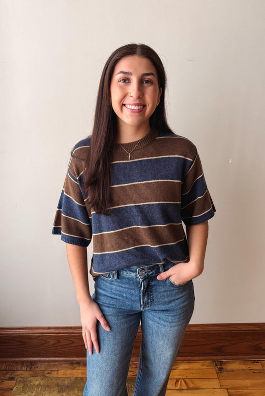 A woman with long dark hair stands indoors wearing a short-sleeve knit top with horizontal brown, navy, and beige stripes, paired with blue jeans, against a plain off-white wall.
