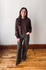 A woman stands in a room with a light gray wall and wooden floor, wearing a dark brown long-sleeve knit sweater and dark gray wide-leg pants.