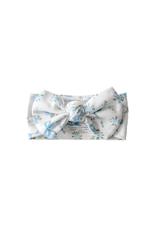 A white fabric headband with a large bow, featuring a delicate light blue floral pattern and a printed brand label in the center, shown on a plain white background.