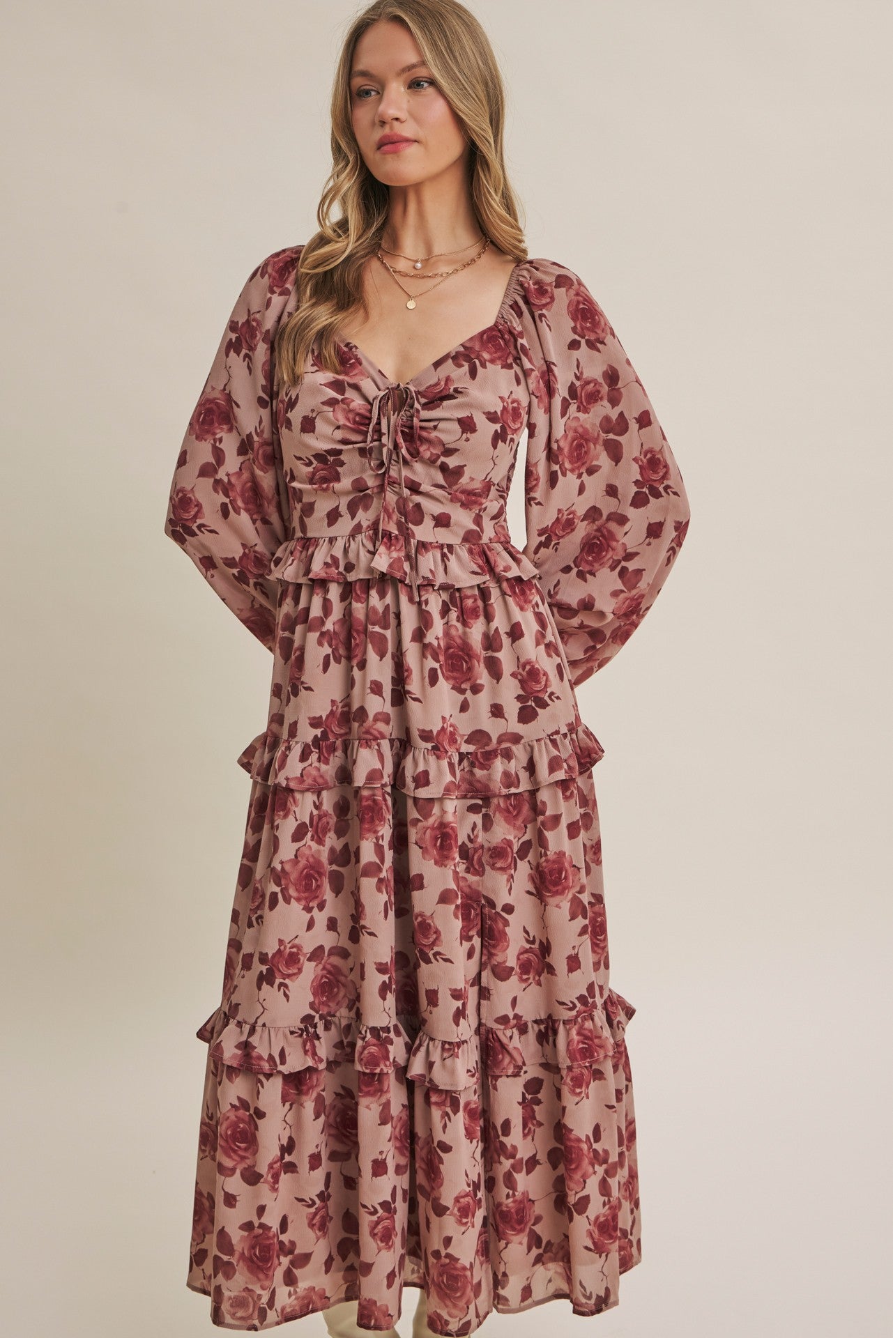 A woman modeling a long-sleeved, tiered maxi dress with a dusty rose and burgundy floral pattern against a solid beige background.