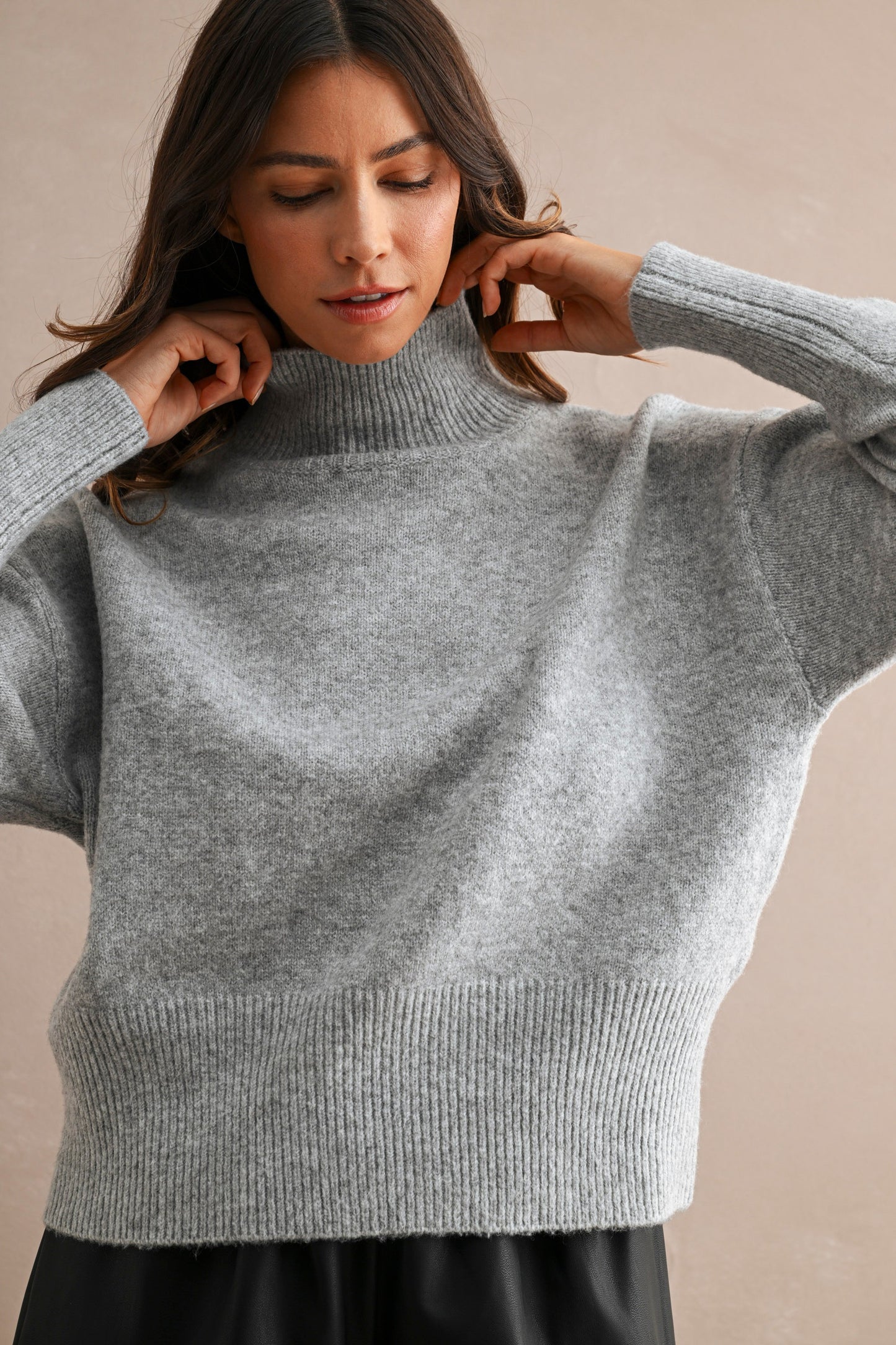 A woman wearing a light gray ribbed knit turtleneck sweater with wide sleeves, posing against a neutral beige background.