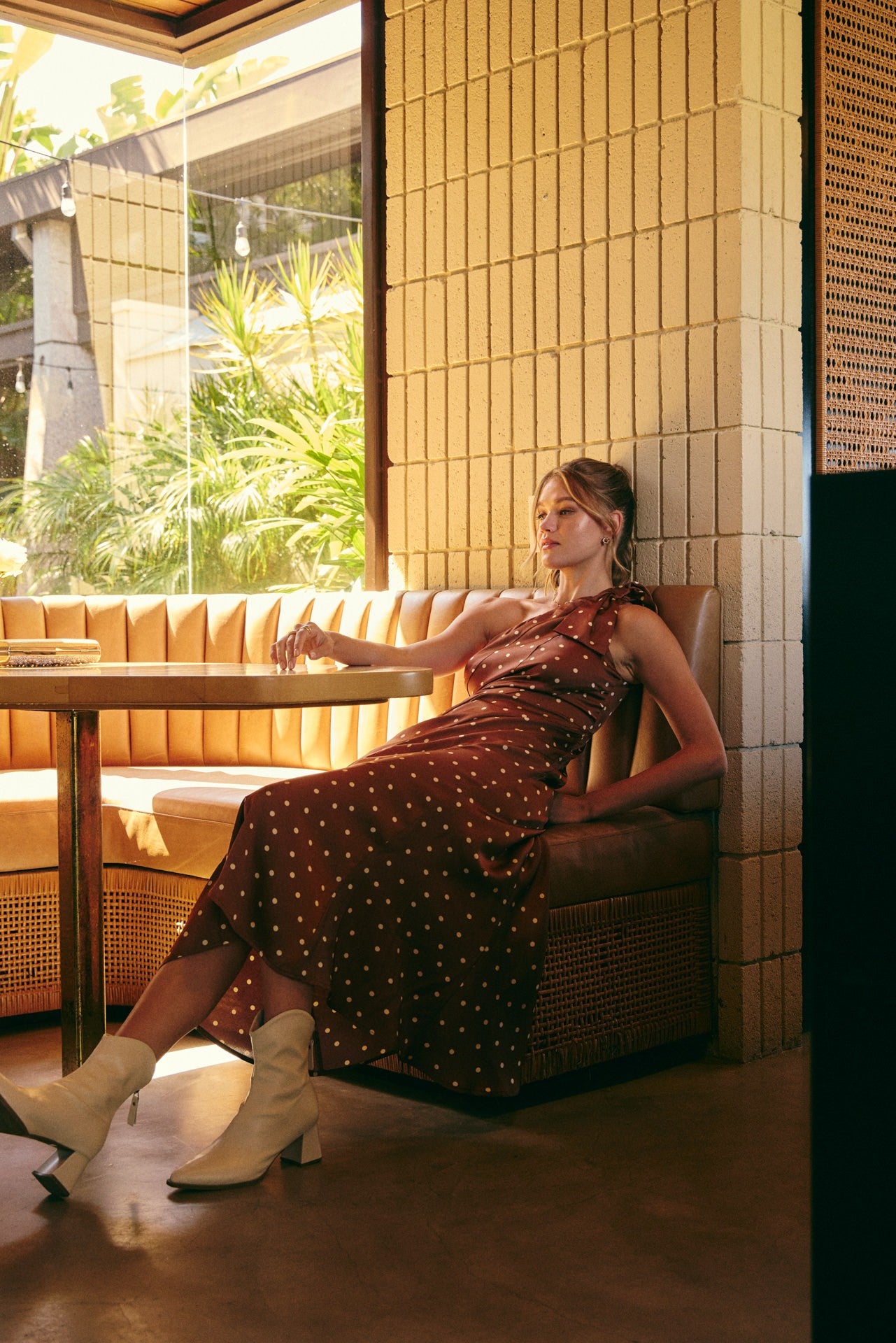A woman wearing a dark brown halter-neck midi dress with white polka dots and white heeled ankle boots, sitting in a tan leather booth inside a modern restaurant with tiled walls.
