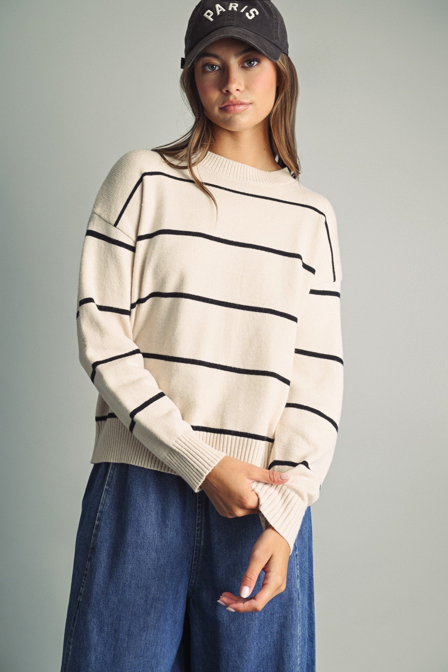 A woman wearing a cream-colored knit sweater with thin black horizontal stripes, paired with blue jeans and a black baseball cap with 'PARIS' embroidered on it, set against a solid gray studio background.