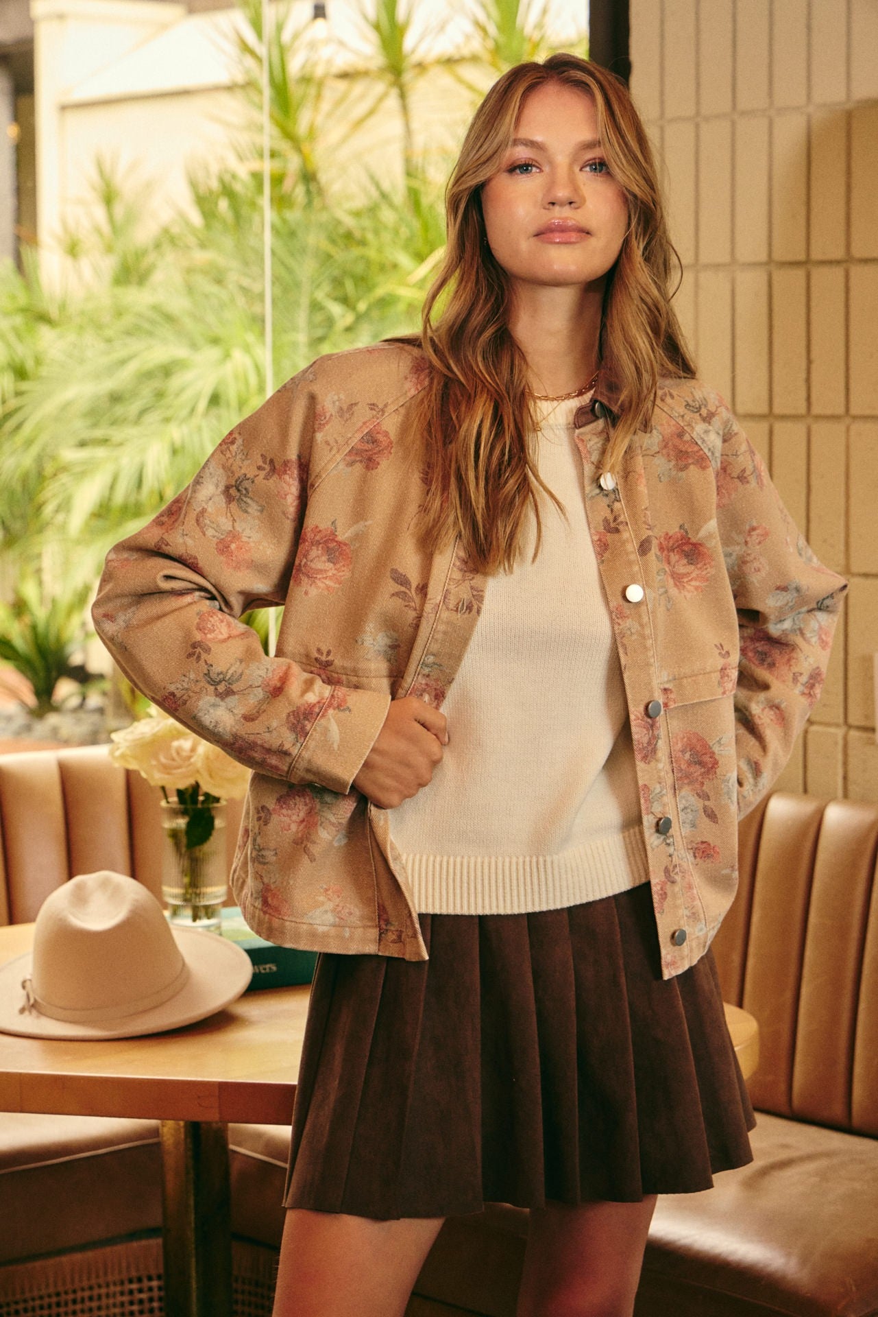 A woman stands in a cafe setting wearing a beige floral button-up jacket over a cream ribbed sweater and a dark brown pleated skirt. A light beige hat rests on a wooden table in the foreground.