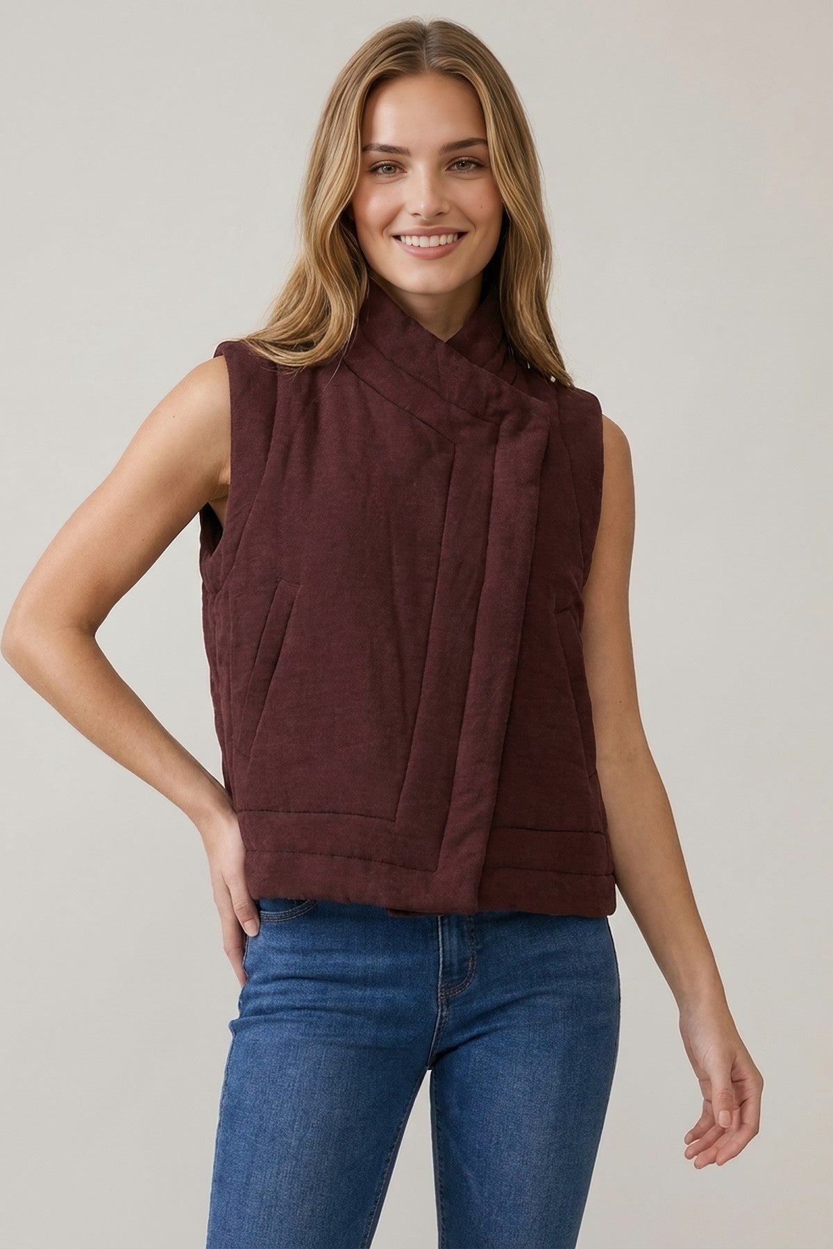 A woman modeling a sleeveless, wrap-style quilted vest in a deep burgundy color, paired with blue jeans, against a plain light gray studio background.