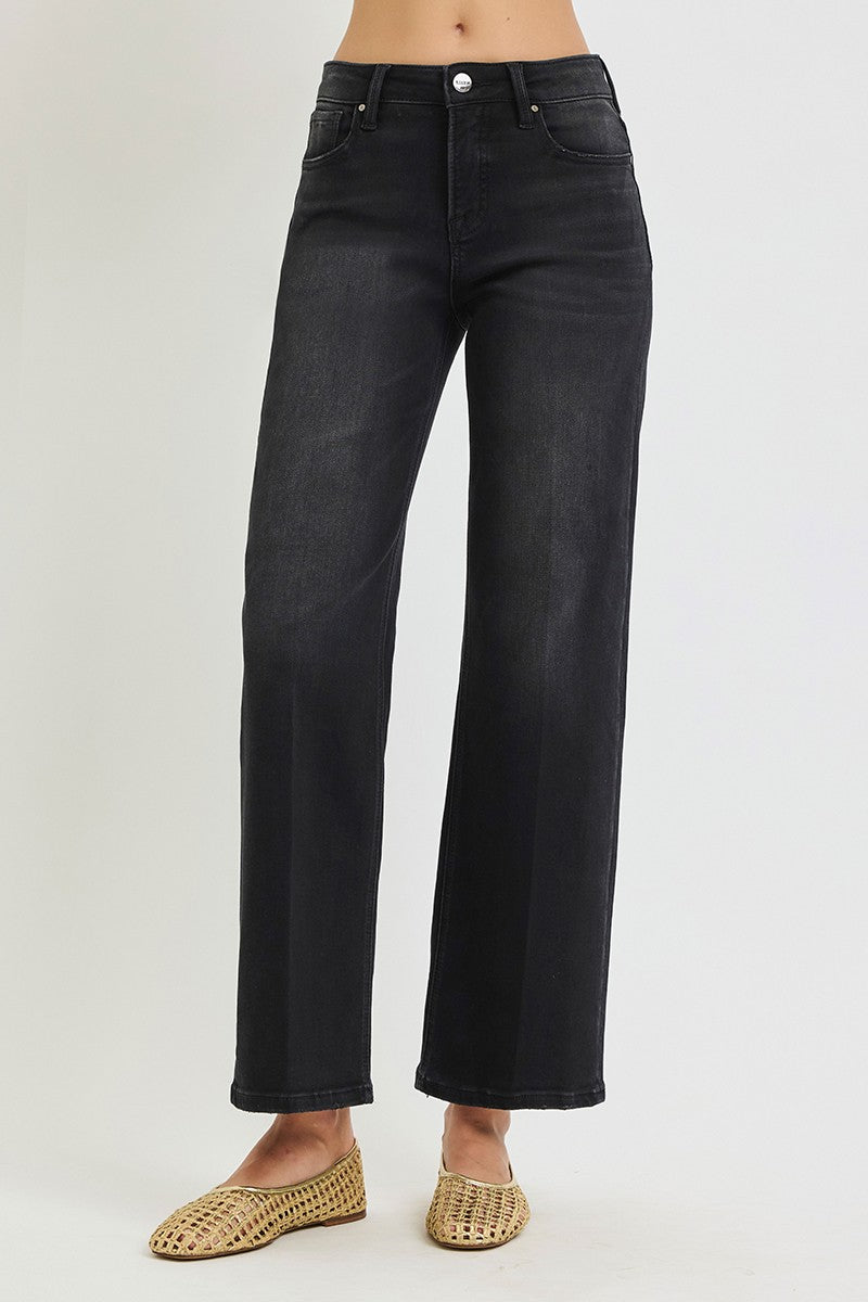 A person wearing dark charcoal black wide-leg jeans and woven tan flat shoes, shown from the waist down against a light gray studio background.
