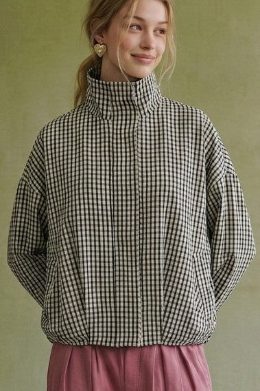 A woman wearing a black and beige gingham patterned quilted jacket with a high mock neck collar, paired with dusty pink trousers, set against a muted olive green background.