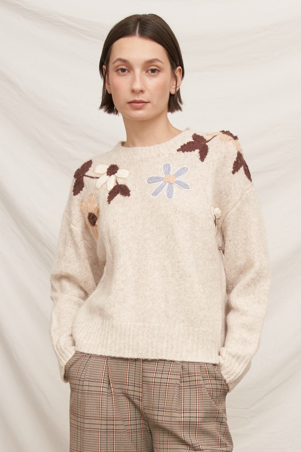 A person wearing a cream knit sweater with brown and beige floral embroidery and a light blue flower motif on the chest, paired with brown plaid trousers, against a soft off-white fabric backdrop.