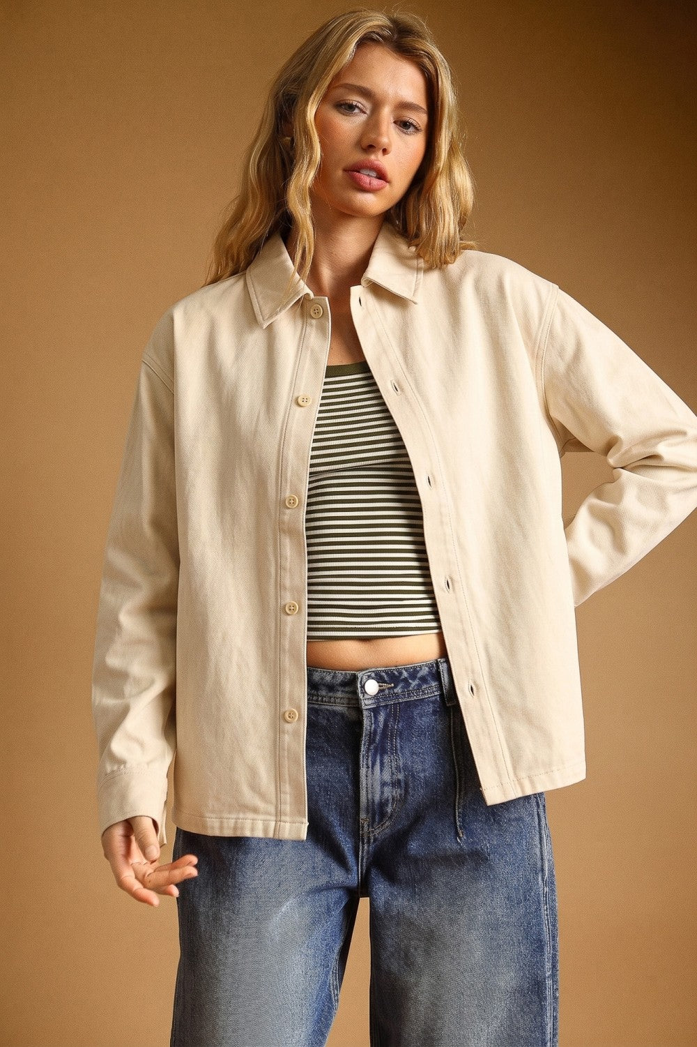 A woman modeling a light beige button-up overshirt over a black and white striped crop top with blue jeans, set against a solid warm brown studio background.