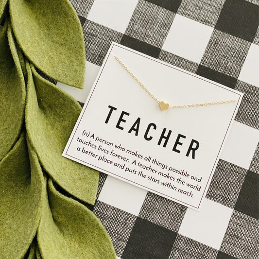 Gold necklace with a heart pendant on a card labeled 'TEACHER' on a checkered background.