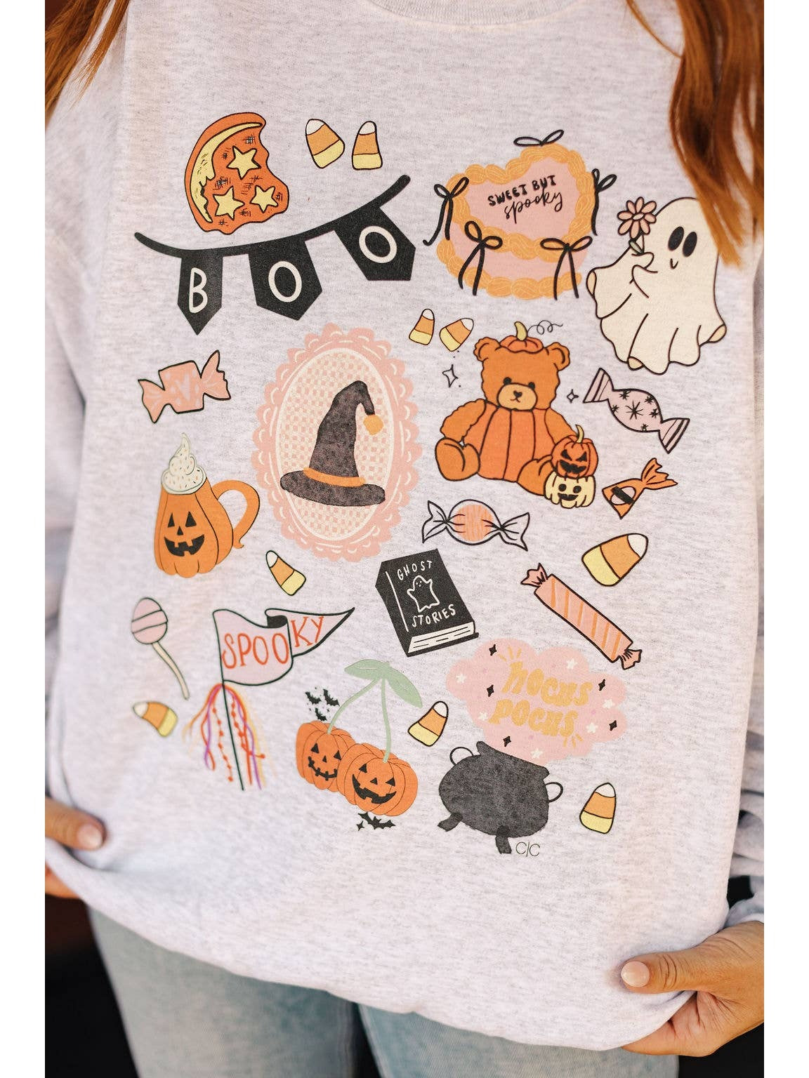 A close-up of a person wearing a light heather gray long-sleeve t-shirt with a colorful Halloween-themed graphic print featuring candy corn, a witch hat, jack-o'-lanterns, a teddy bear, ghosts, and the word 'BOO'.