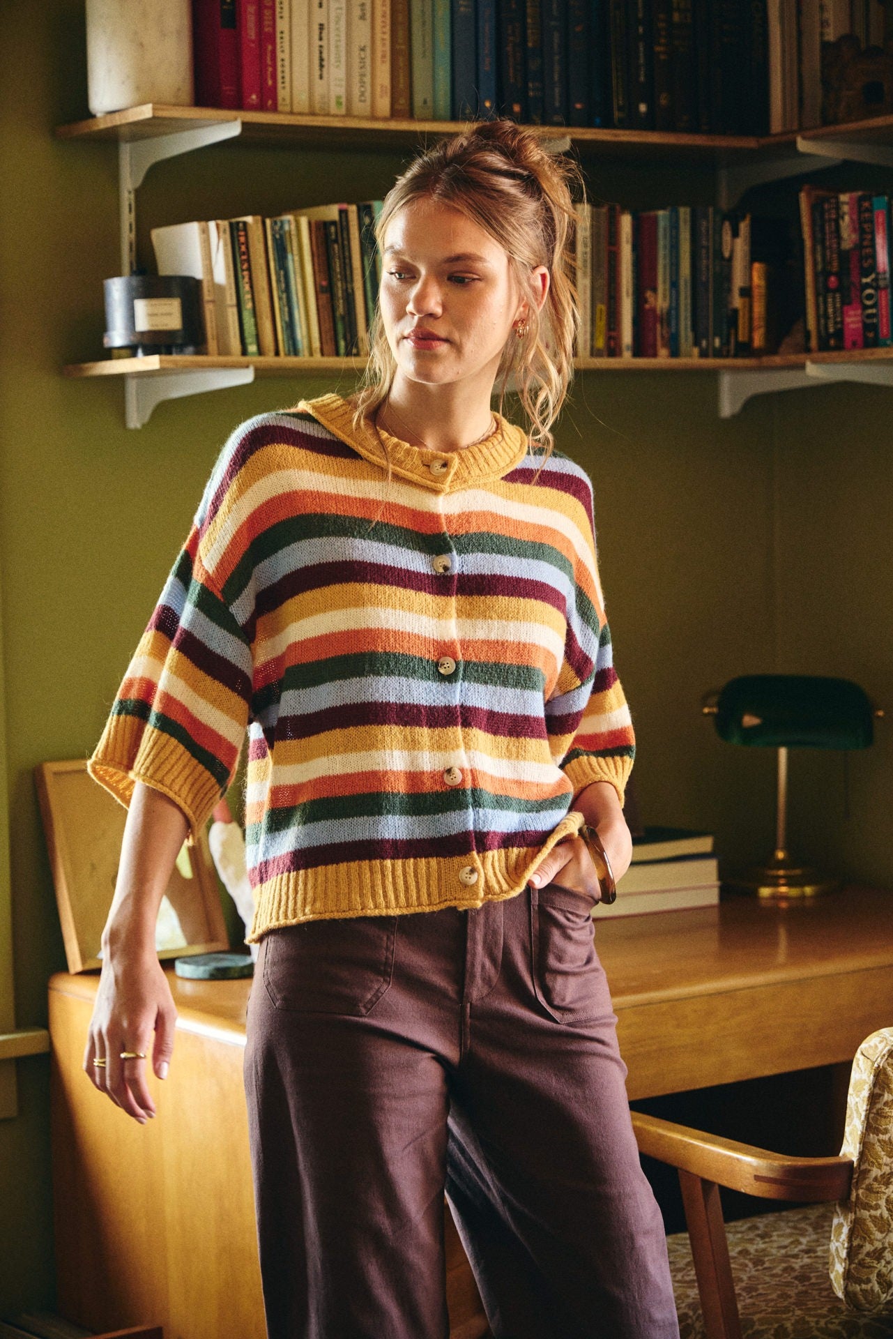 A woman wearing a retro-style striped knit cardigan sweater with yellow, orange, green, blue, and burgundy stripes, paired with dark purple trousers, standing in a cozy room with bookshelves and a desk.