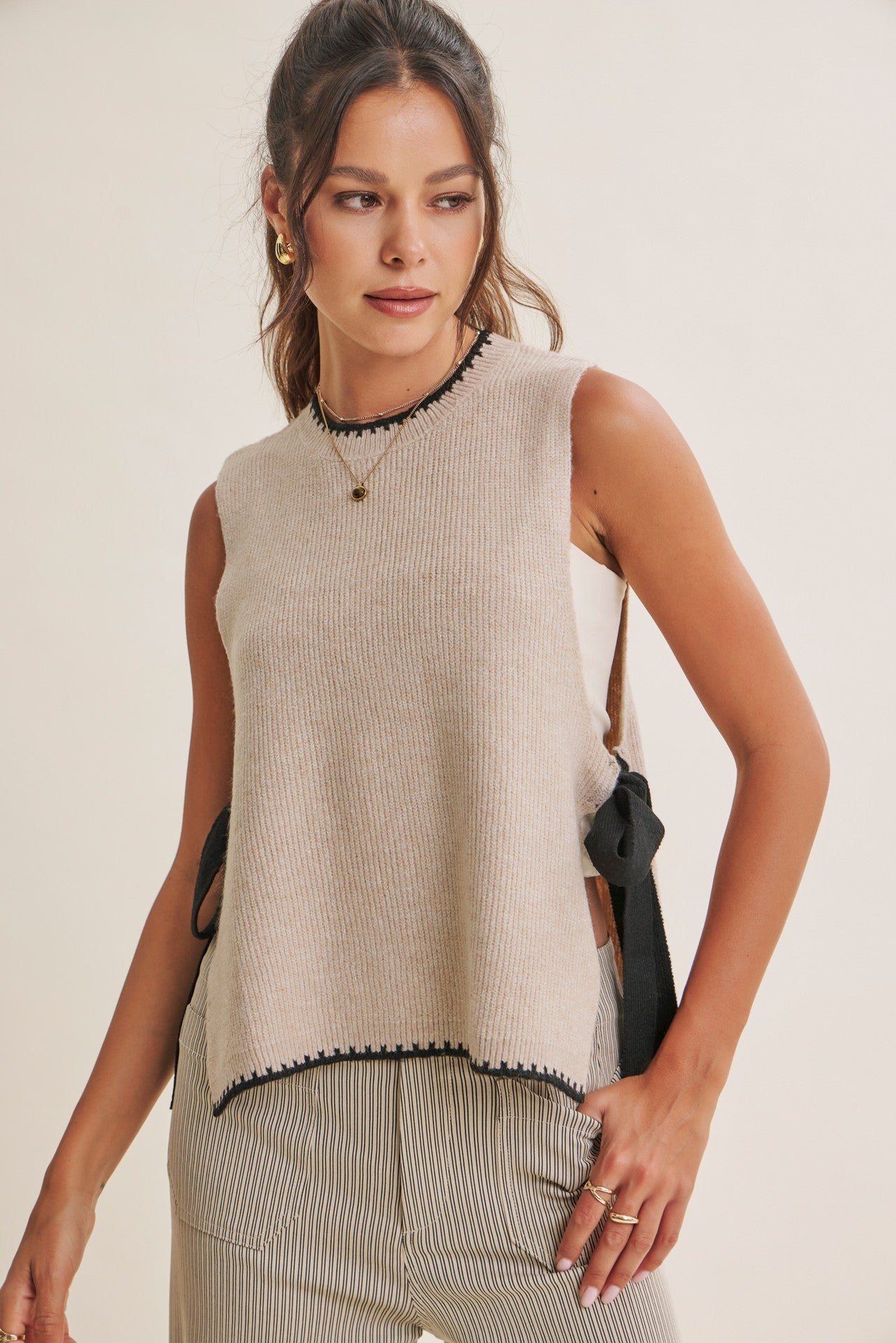 A woman modeling a sleeveless ribbed knit top in a light beige/cream color with black trim details along the neckline and hem, paired with striped pants against a plain light background.