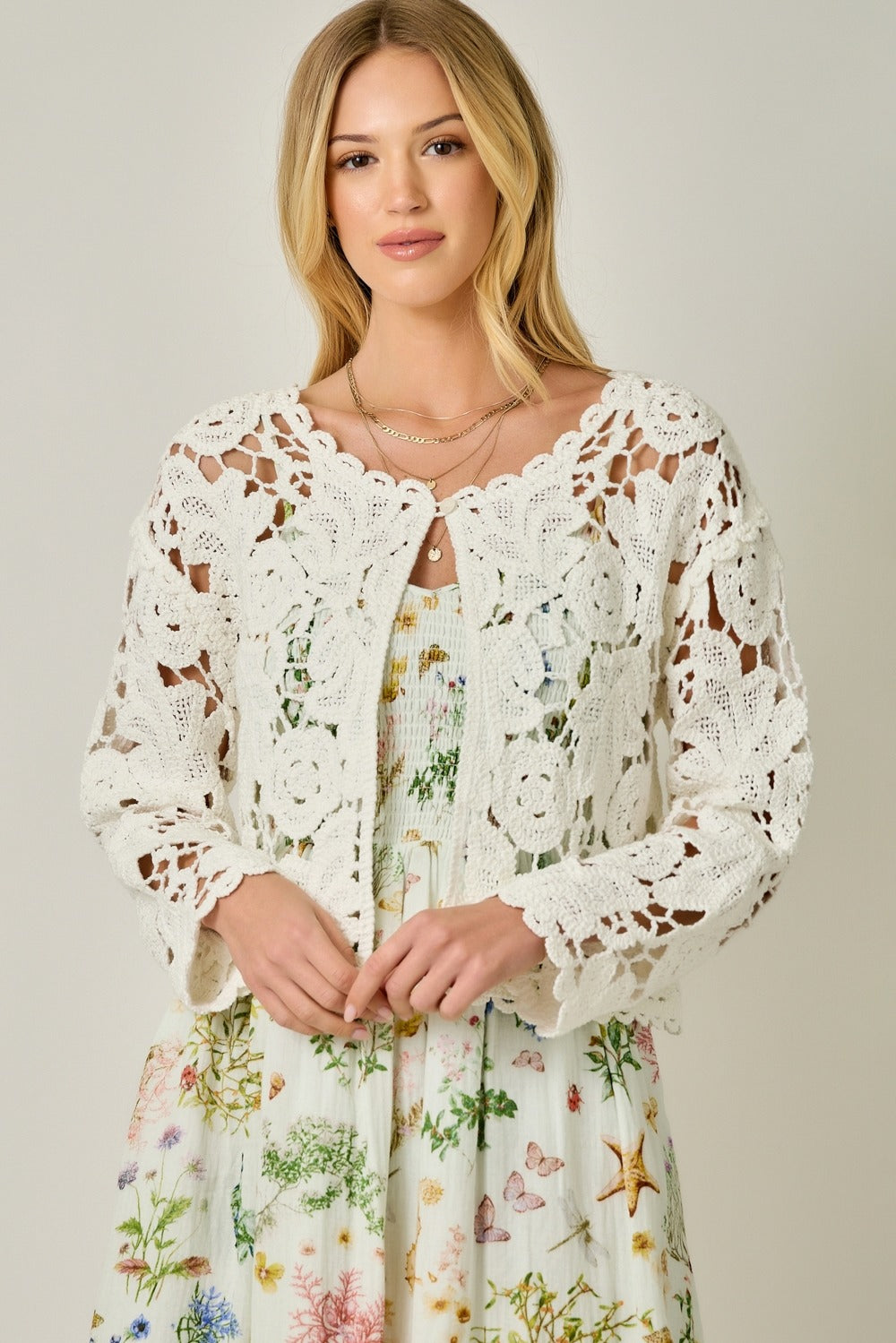 Woman wearing a white lace cardigan over a floral dress on a plain background