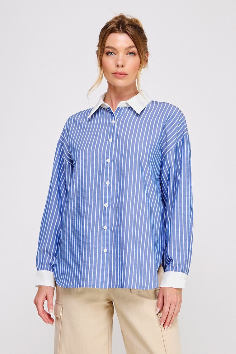 A woman modeling a blue and white vertically striped button-down shirt with a solid white collar and cuffs, paired with beige pants, against a light gray studio background.
