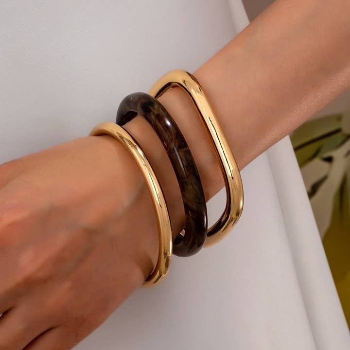 Three bangle bracelets worn on a person's wrist: two slim gold-tone metal bangles and one wider dark brown marbled bangle.