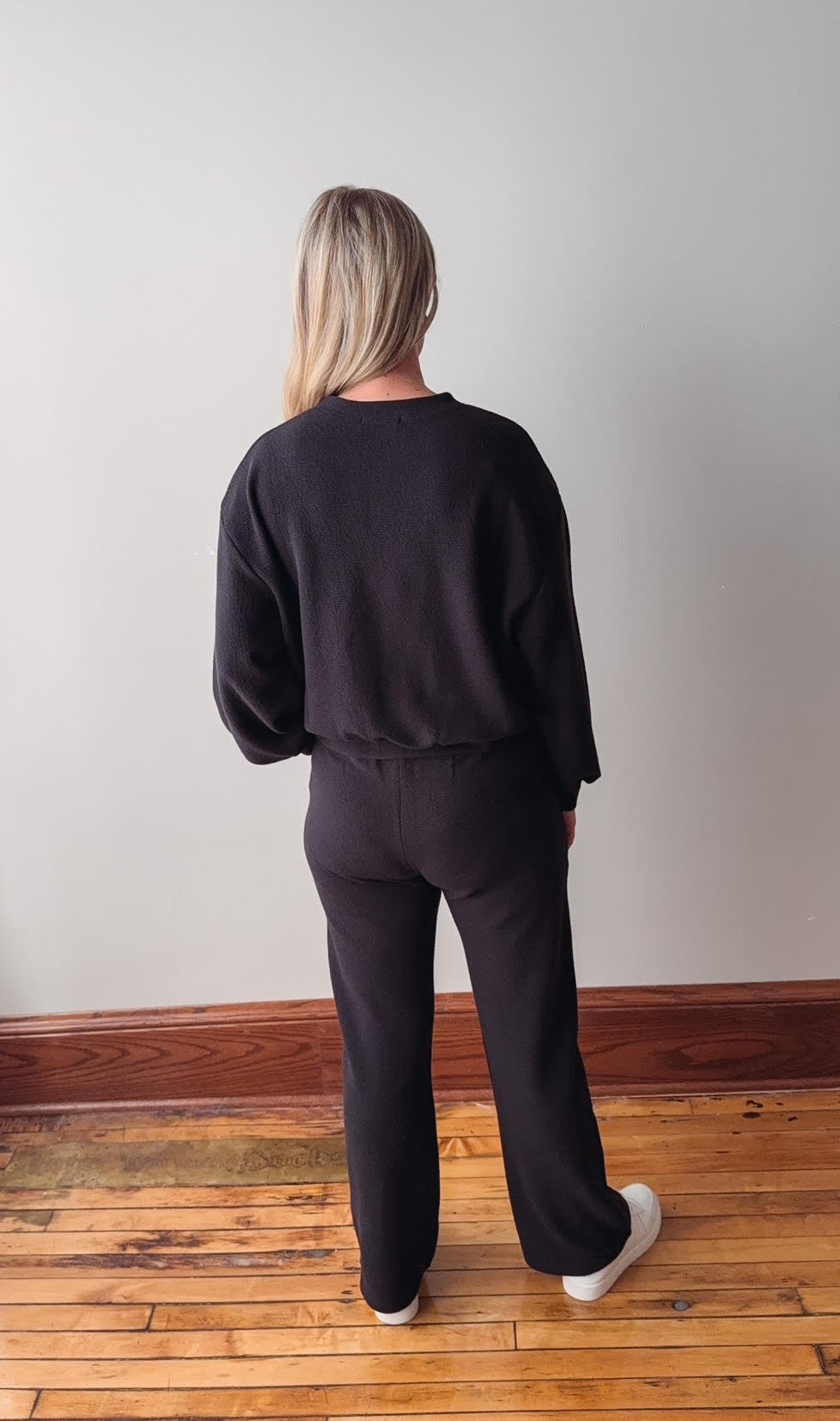 Person wearing a black outfit standing against a plain wall on a wooden floor.