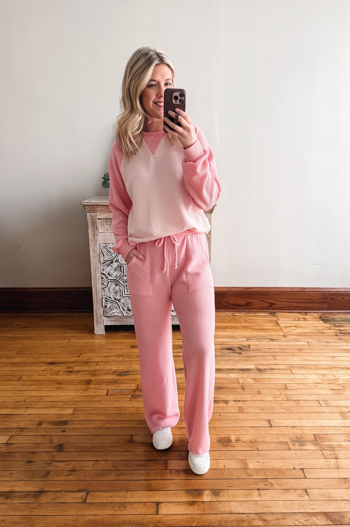 Woman taking a mirror selfie wearing a pink outfit in a room with wooden flooring.
