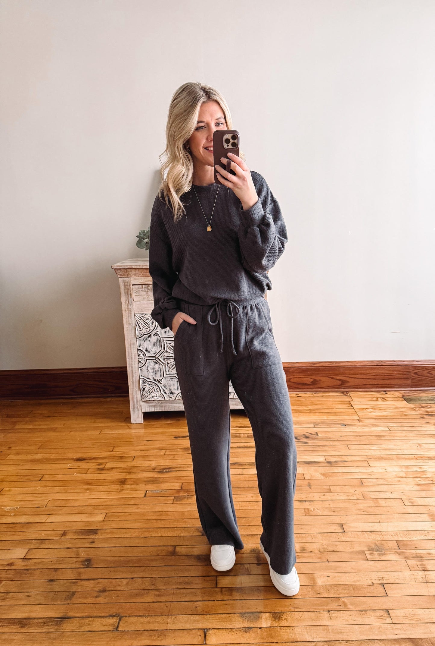 Woman taking a mirror selfie wearing a dark gray sweatshirt and pants in a room with wooden flooring.