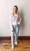 Woman wearing a white sweatshirt with a graphic design, light blue jeans, and white sneakers, standing against a plain wall.