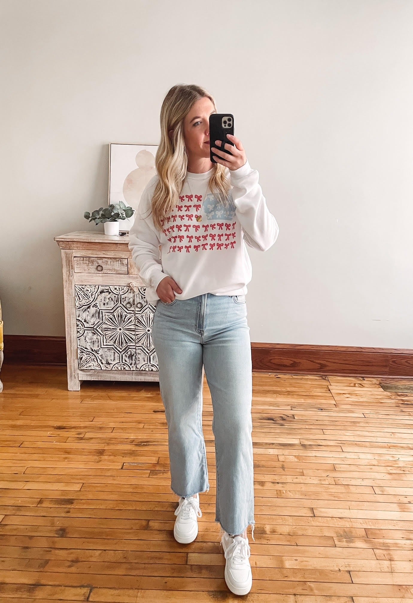 Person taking a mirror selfie wearing a white sweatshirt with a colorful pattern and light blue jeans.