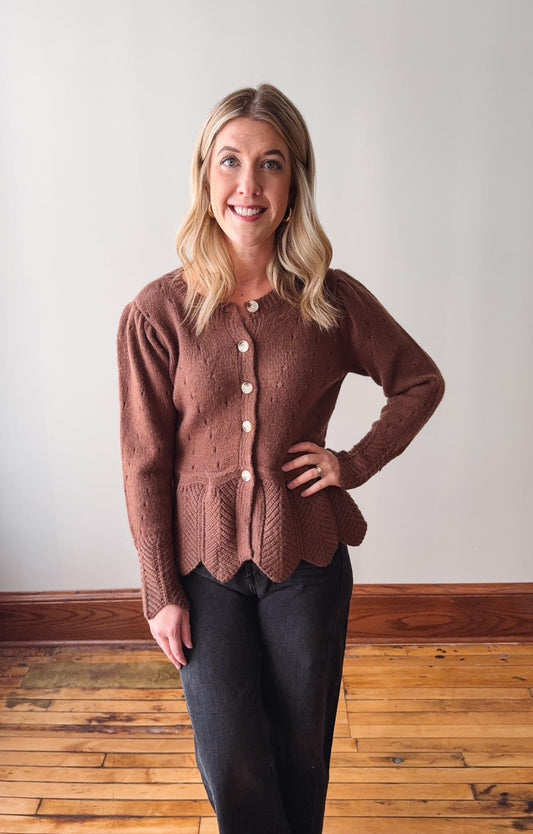 Woman wearing a brown knitted cardigan standing on a wooden floor with a white wall background