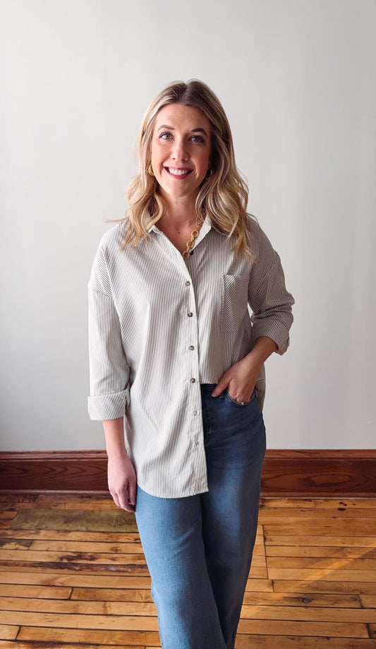 Woman wearing a beige button-up shirt and blue jeans standing in a room with wooden flooring and a white wall.