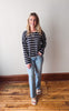 Woman wearing a striped sweater and jeans standing on a wooden floor with a white wall background