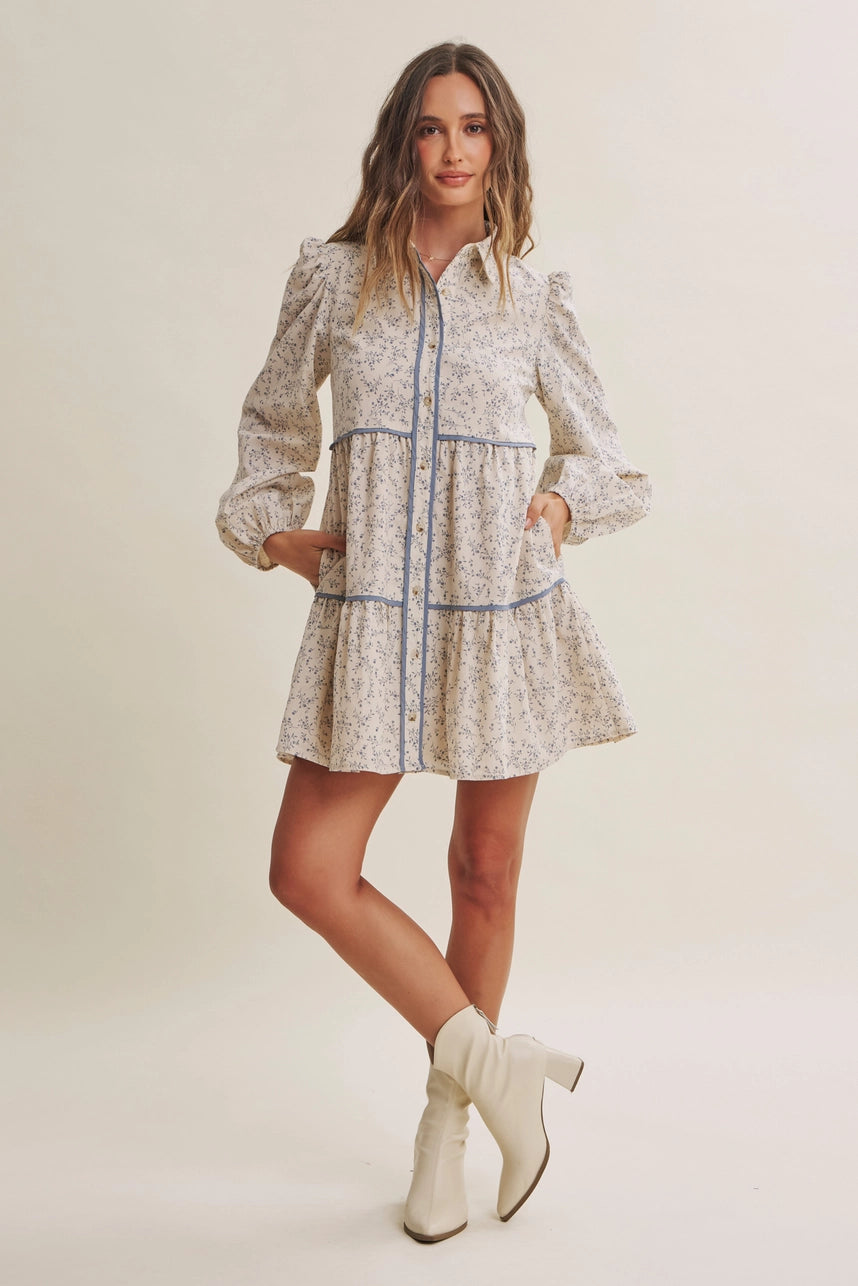 A woman modeling a long-sleeve mini dress with a beige and white floral print, blue piping details, and cream-colored ankle boots, set against a plain beige studio background.
